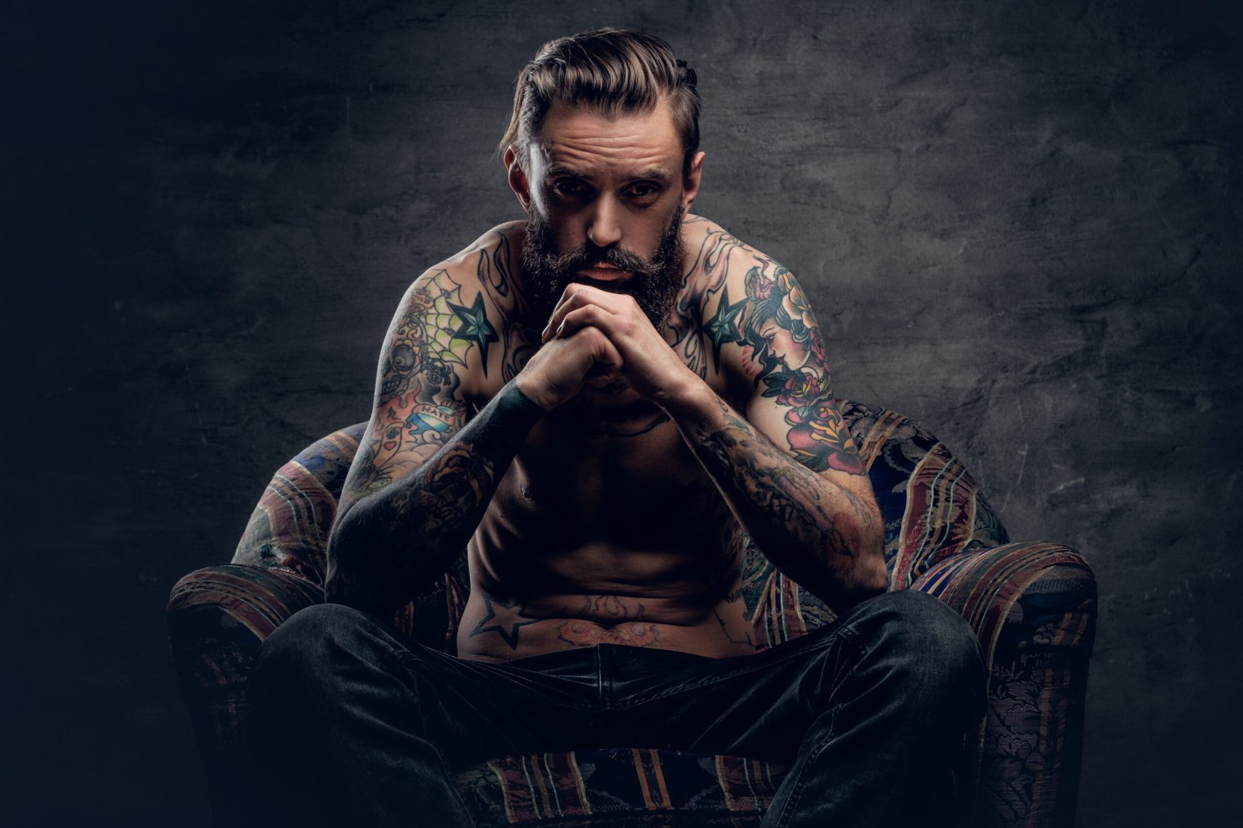Portrait of a thoughtful bearded male with tattooed naked torso and crossed arms, sits on a chair over dark grey background.